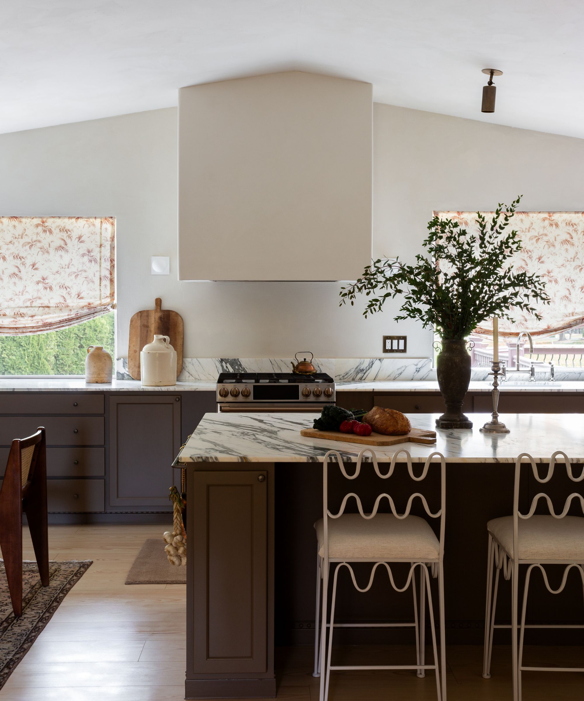 Kitchen with broccoli brown by Farrow &amp;amp; Ball painted island and cabinets, white marble countertops, wrought-iron wiggly white bar stools, a large oven hood, and floral cream blinds.