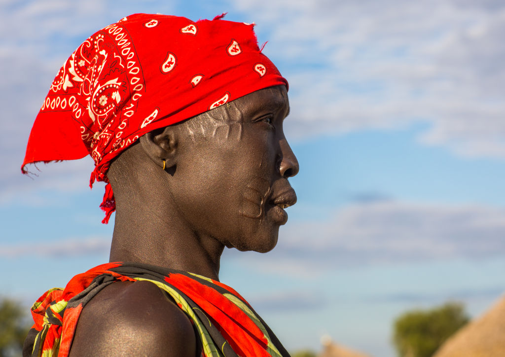 A dark skinned person wearing a red bandana and a red and yellow tank top looks to the right, their face lined with light colored scars.