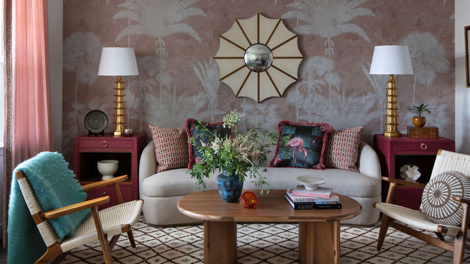 Vibrant living room with pink-and-white 'tree' wallpaper with a mirror and pink curtains. In the room, a sofa, two armchairs, a wooden coffee table, two side tables with lamps, and a patterned rug are pictured.
