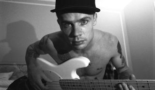 Musician Flea of the Red Hot Chili Peppers poses shirtless in his hotel room for a portrait, while holding his guitar and wearing a baseball cap