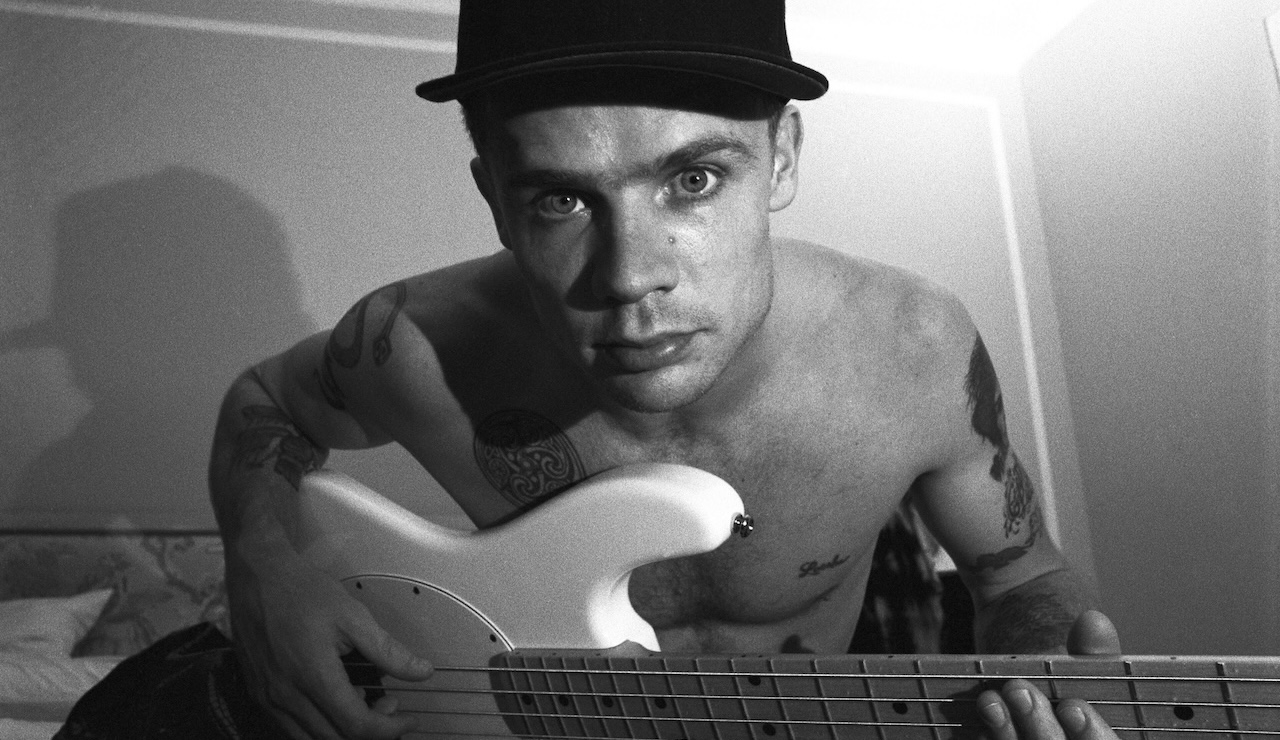 Musician Flea of the Red Hot Chili Peppers poses shirtless in his hotel room for a portrait, while holding his guitar and wearing a baseball cap