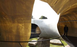 Interior and exterior view of the 2014 Serpentine Pavilion from inside. The grass and stones below along with the sky can be seen through an opening in the structure and there is a person standing inside