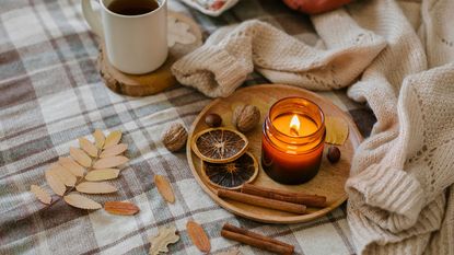 A round wooden tray with some dried orange slices and a lit candle on it, on top of a white surface with plaid material off cuts, dried autumnal leaves, and some cinnamon sticks 