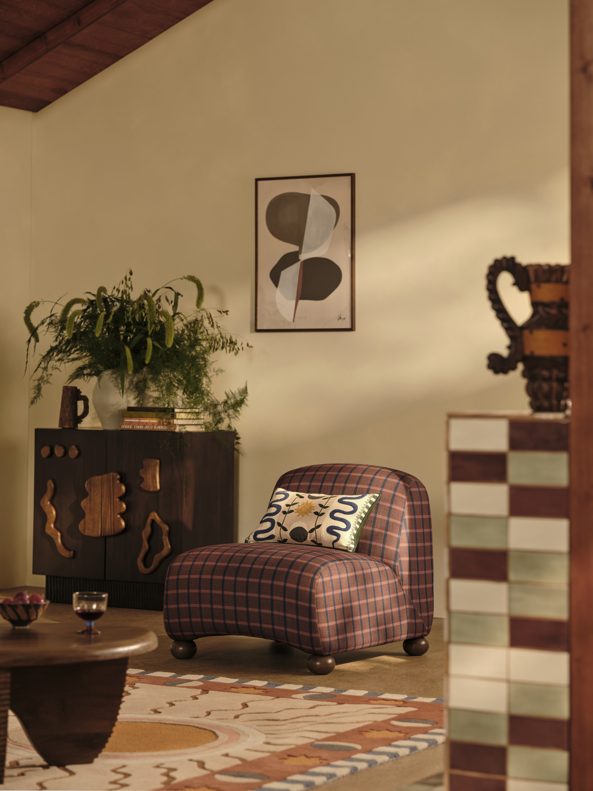 Image of a modern living room with a purple, plaid accent chair that has wooden ball feet. There is a wooden coffee table in front of it and a wooden cabinet with abstract handles behind it.