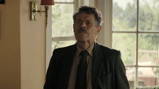 Willem Dafoe wearing a suit and tie during Tenzing's trailer.