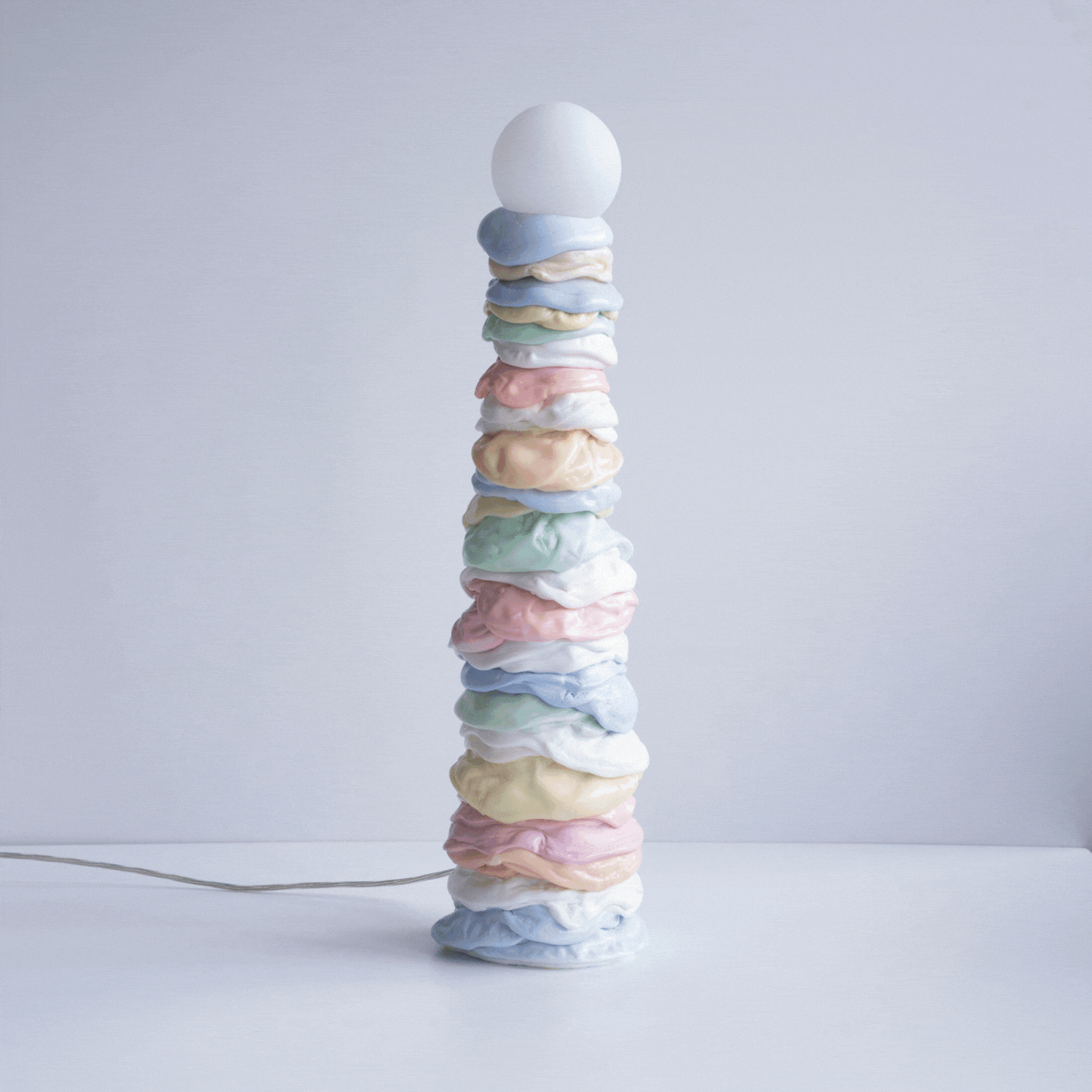 A squishy-looking lamp made of pastel-shaded rings of melted plastic, with a round light bulb at its peak, sits in a white room while lighting on and off intermittently.