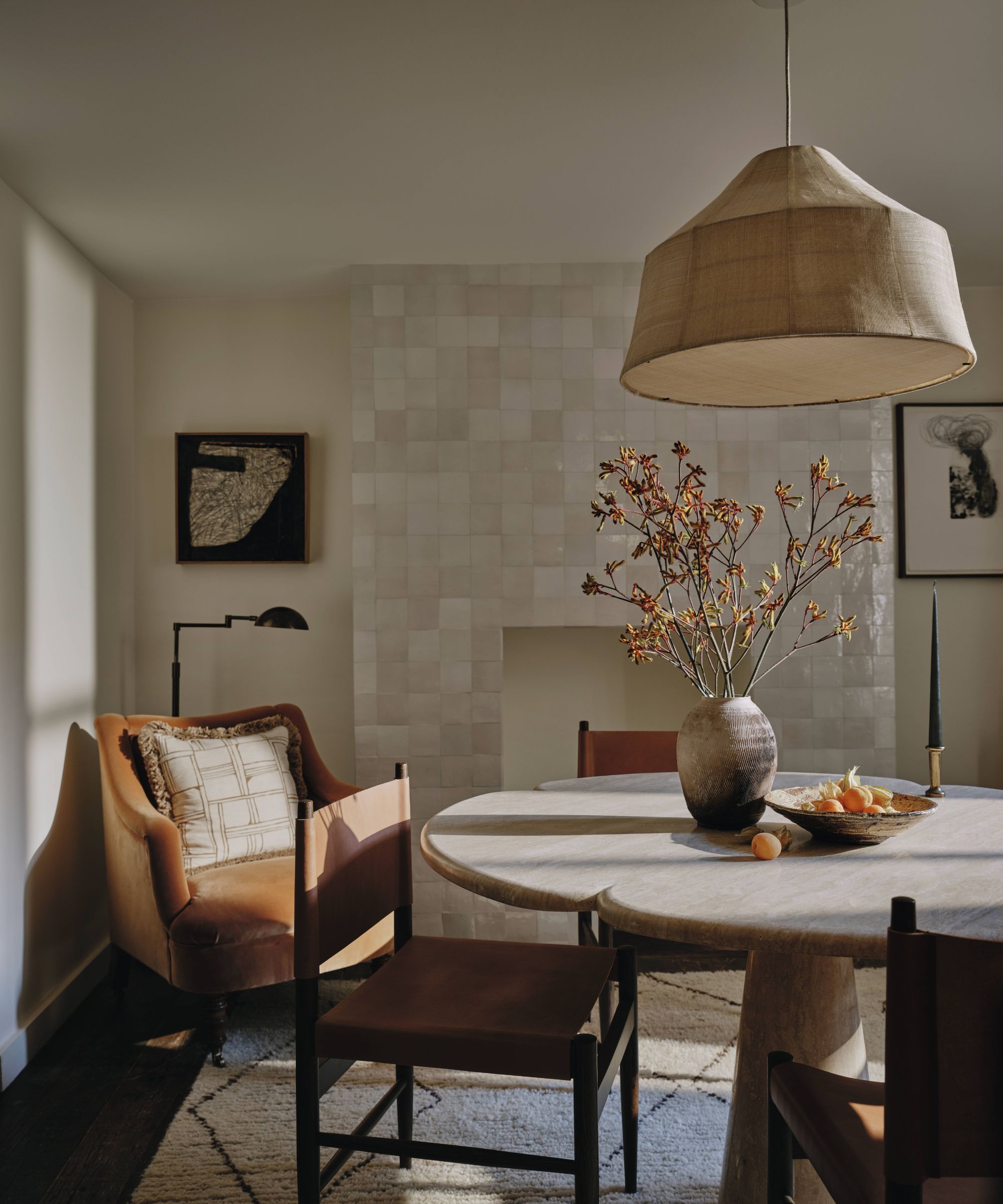 A photograph of a mid-century modern dining room featuring a round stone table, four wooden chairs with leather sling seats, and a textured beige-colored pendant lamp.