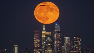 the new york city skyline below and a huge supermoon shining above, it is slightly colored orange. 