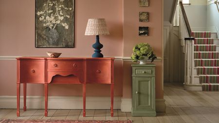 A pink hallway in a grand house with a pink painted wooden dressing table with a blue lamp on top, and a green painted side cabinet next to it with a vase of green flowers on top. 