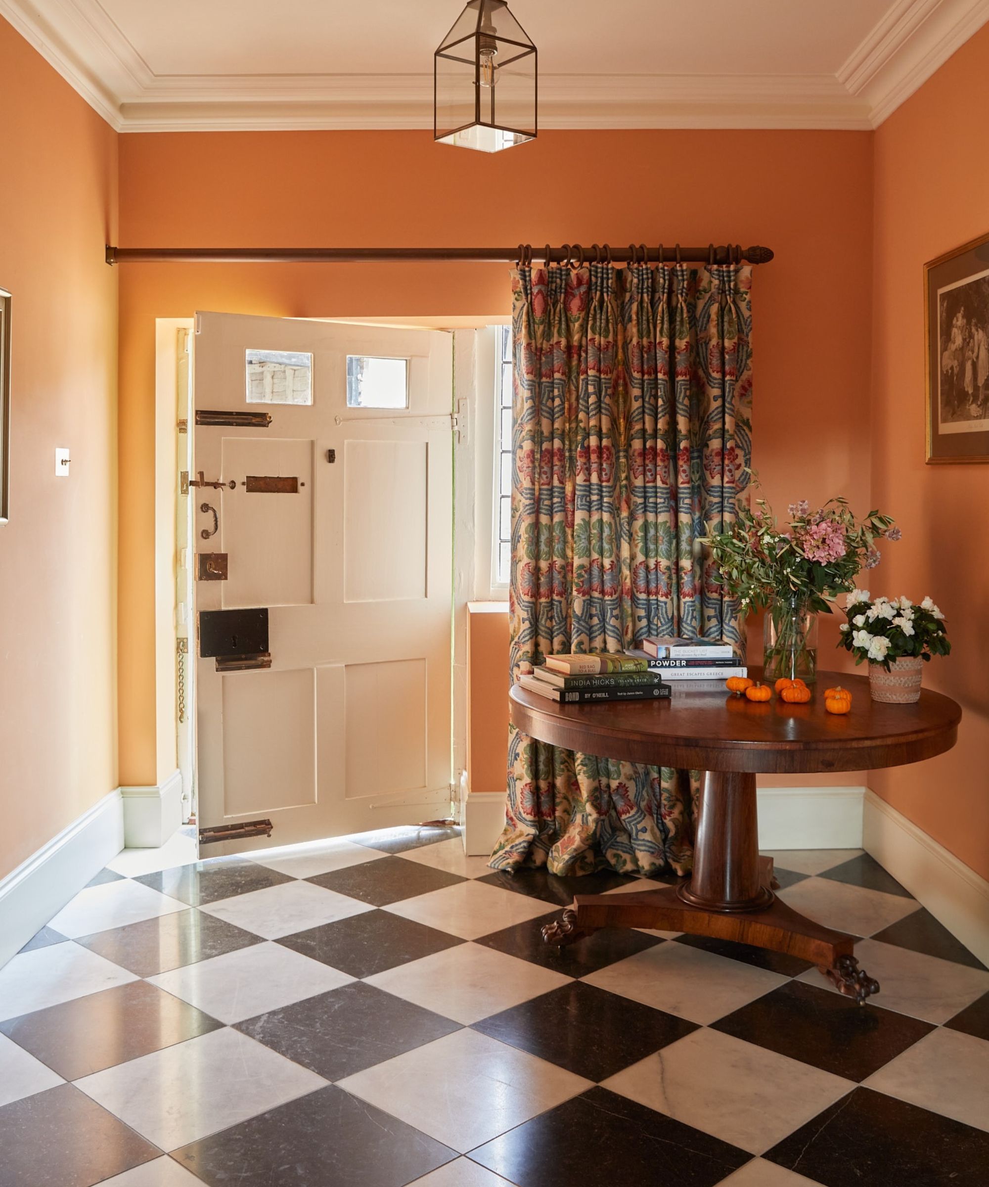 Entryway with orange painted walls, black and white tiled floor, a large wooden round table with books and vases of flowers on top, a large white front door with a heavy floral curtain beside it