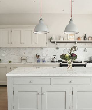 A kitchen with a light gray marble kitchen island with a glass vase of flowers on it, white kitchen cabinets behind it, and three gray pendant lights hanging from the ceiling