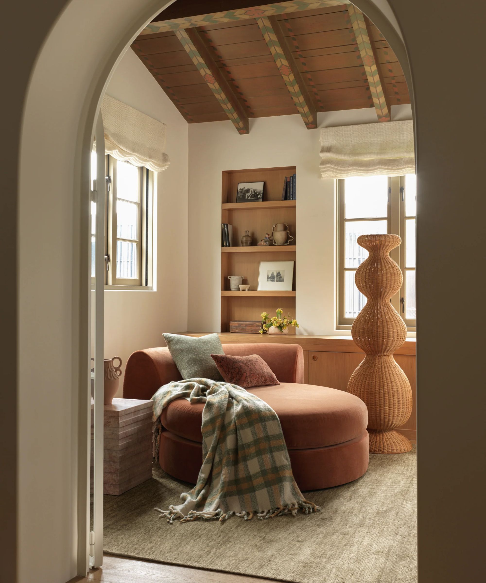 a sitting room with a large, round chair in soft pink velvet, pillows, a blanket draped across the side, a large rattan sculpture, built in shelving, a solid rug, and painted ceiling beams