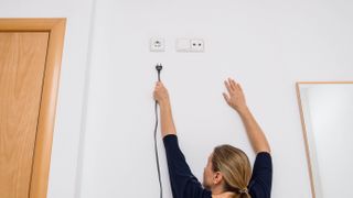 A person reaching for a plug outlet which is high up the wall