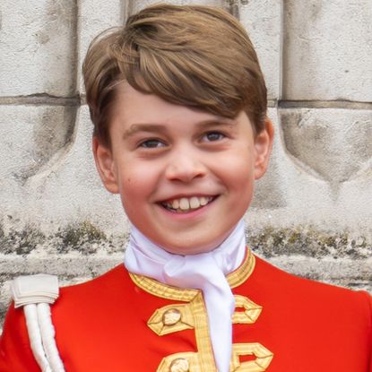 Prince George in a red and gold uniform smiling in front of a stone pillar