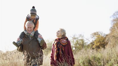 An older man smiles and carries a child on his shoulders while he and an older woman walk through a field in the fall.