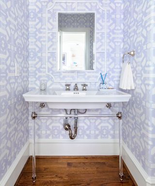 A small bathroom with lilac and white patterned wallpaper with a wall sconce with three lampshades, a rectangular mirror, and a white freestanding sink