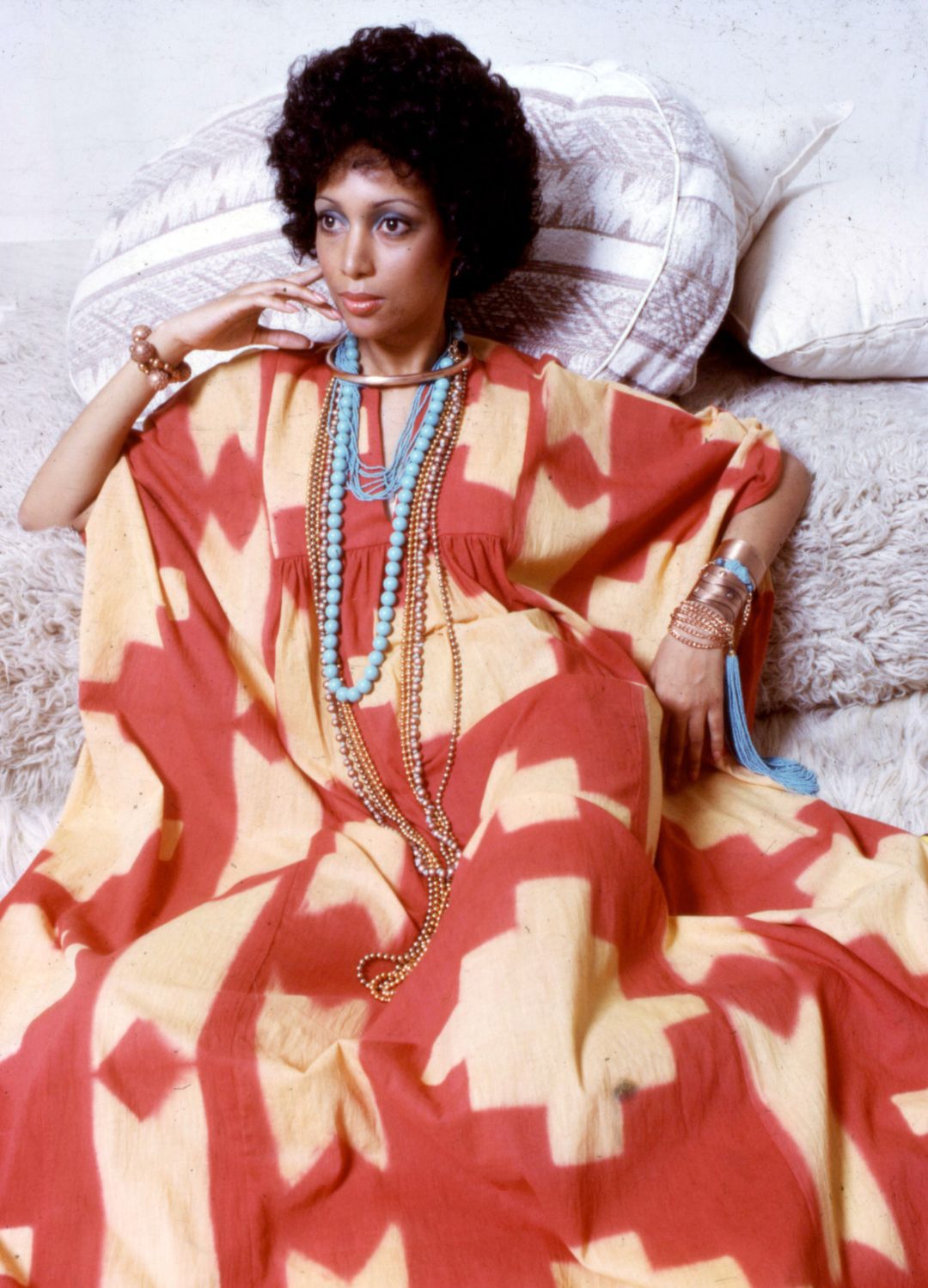 Portrait of an unidentified model, in a red and yellow print dress accented with gold and turquoise jewelry, as she poses on a set of neutral-colored throw pillows, New York, 1970s.