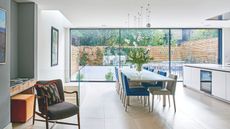 A bright kitchen with beige tiles and large windows, a white island and a long dining table and chairs in blue and grey tones. To the left is a green wall and a desk, with a brown wooden chair and patterned cushion. Behind, through the windows, a lush and leafy garden can be seen.