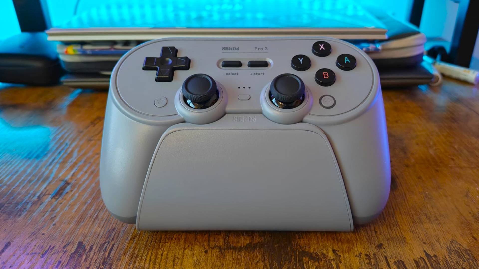 8BitDo Pro 3 controller on its charging stand sitting on woodgrain desk.