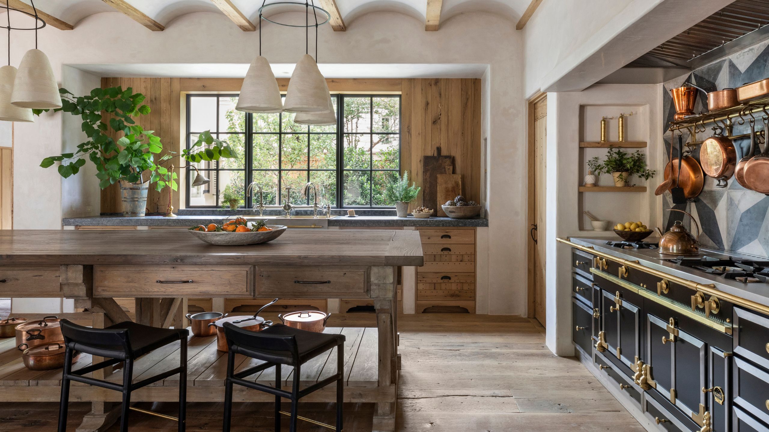 Rustic kitchen with a wooden island, black bar stools, a black and brass range stove, hanging copper pots, and large windows bringing in natural light over the sink.