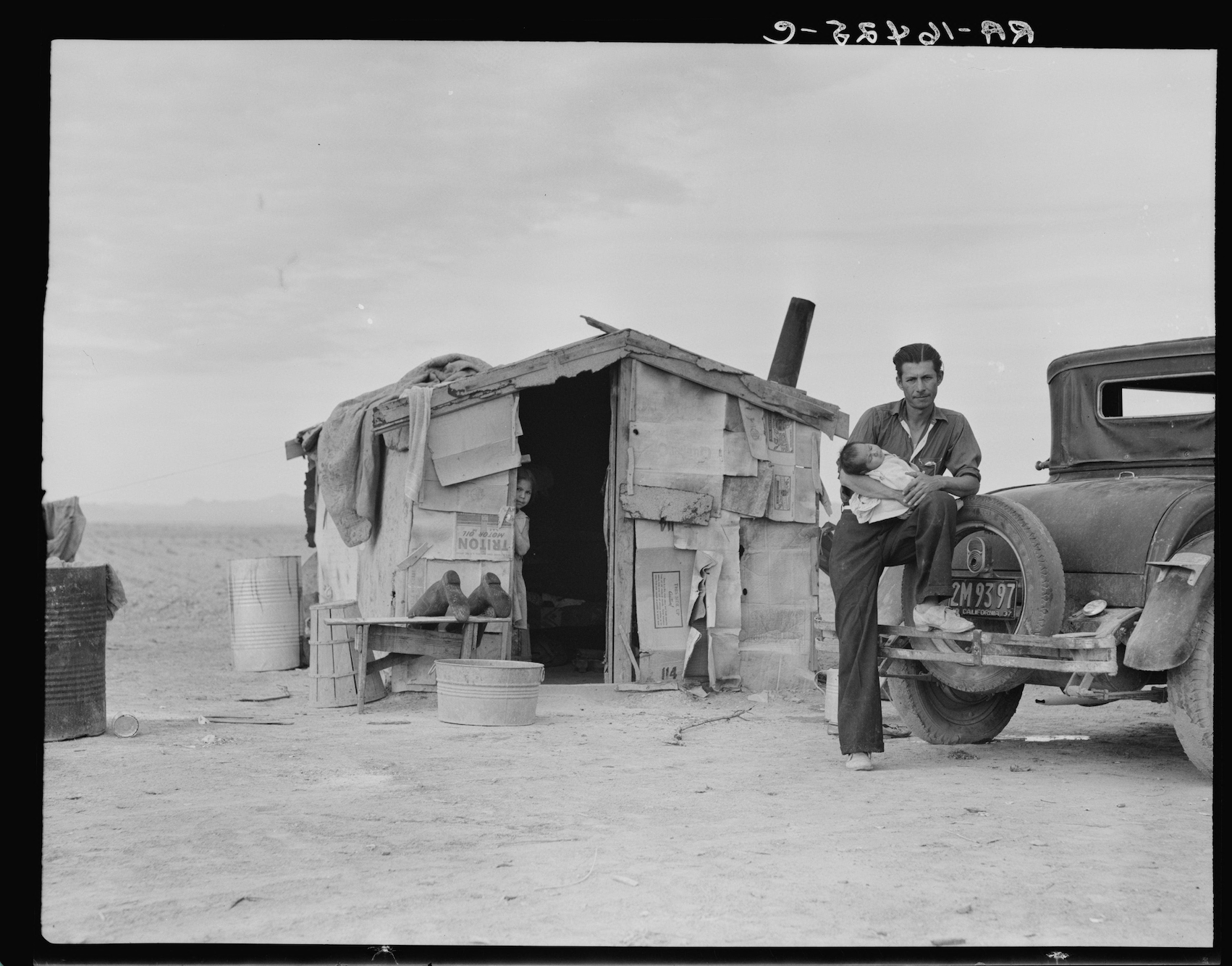 A man holding a sleeping infant leans against an old car beside a crude shack made of scrap wood and cardboard, with a young girl standing in the doorway, all on bare, dusty ground.