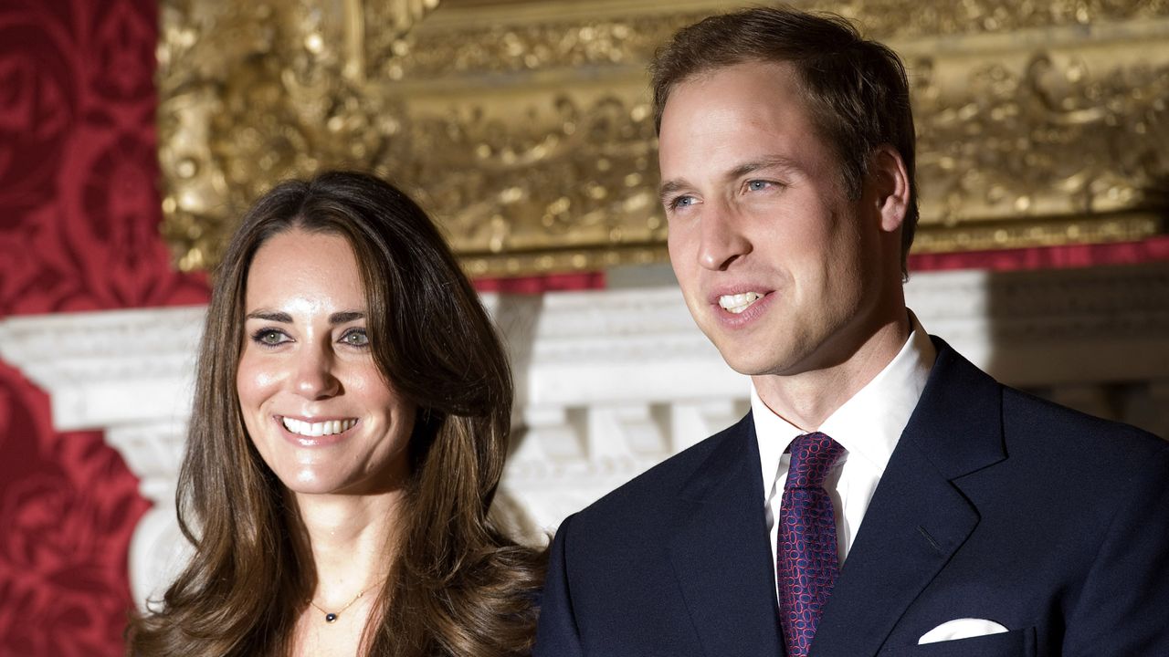 Kate Middleton wearing a blue dress and Prince William standing in front of a red wall and gold painting wearing a suit and tie