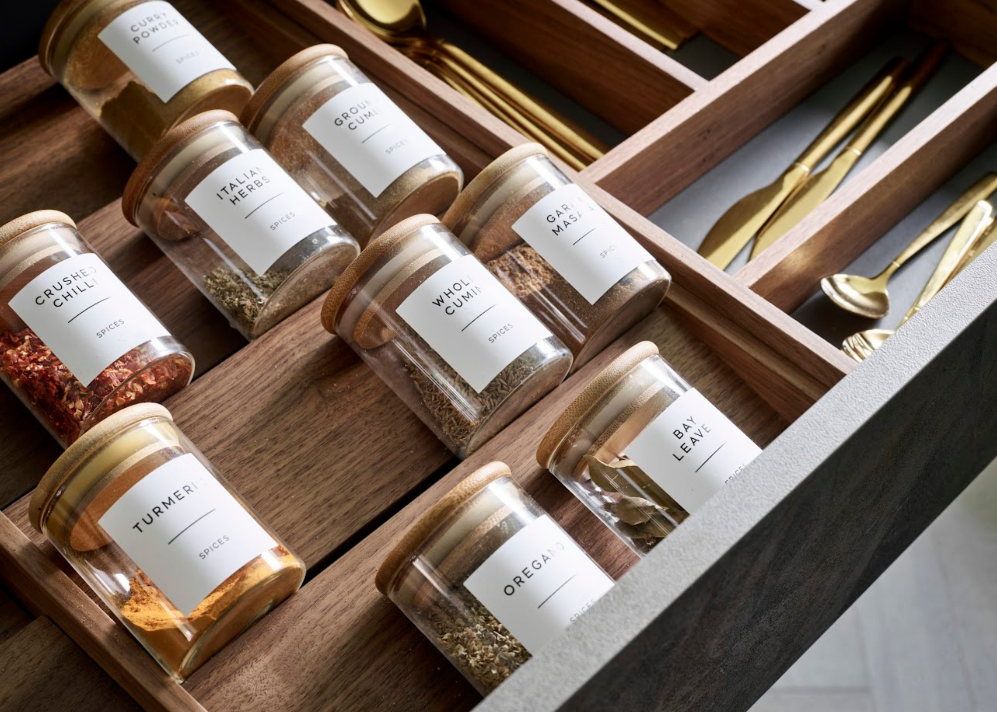A close up of inside a wooden kitchen drawer with a spice rack insert, uniformed labelled jars and gold cutlery in the background