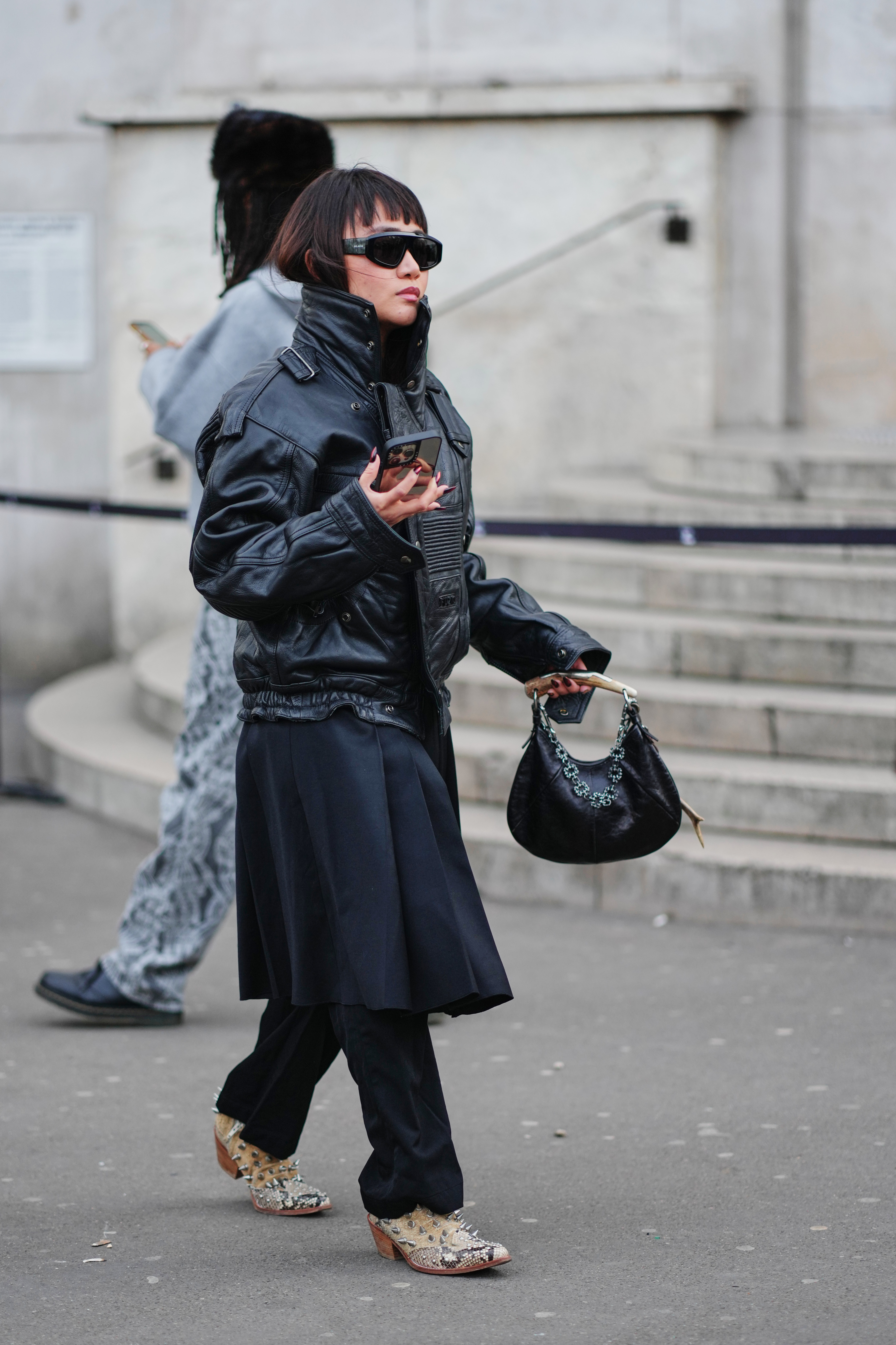 PARIS, FRANCE - JANUARY 20: A guest wears a straight black bob haircut with short bangs, black wraparound sunglasses, a black leather shoulder bag with a short metal handle and a silver metal chain detail, a black leather bomber jacket with a high stand collar, snap buttons, shoulder epaulettes, zip and flap pockets and a ribbed hem, a black pleated skirt, black trousers, beige straw woven pointed-toe cowboy boots shoes with silver metal stud embellishments and brown stacked heels, outside 424, during Paris Fashion Week - Menswear Fall/Winter 2026-2027, on January 20, 2026 in Paris, France (Photo by Edward Berthelot/Getty Images)