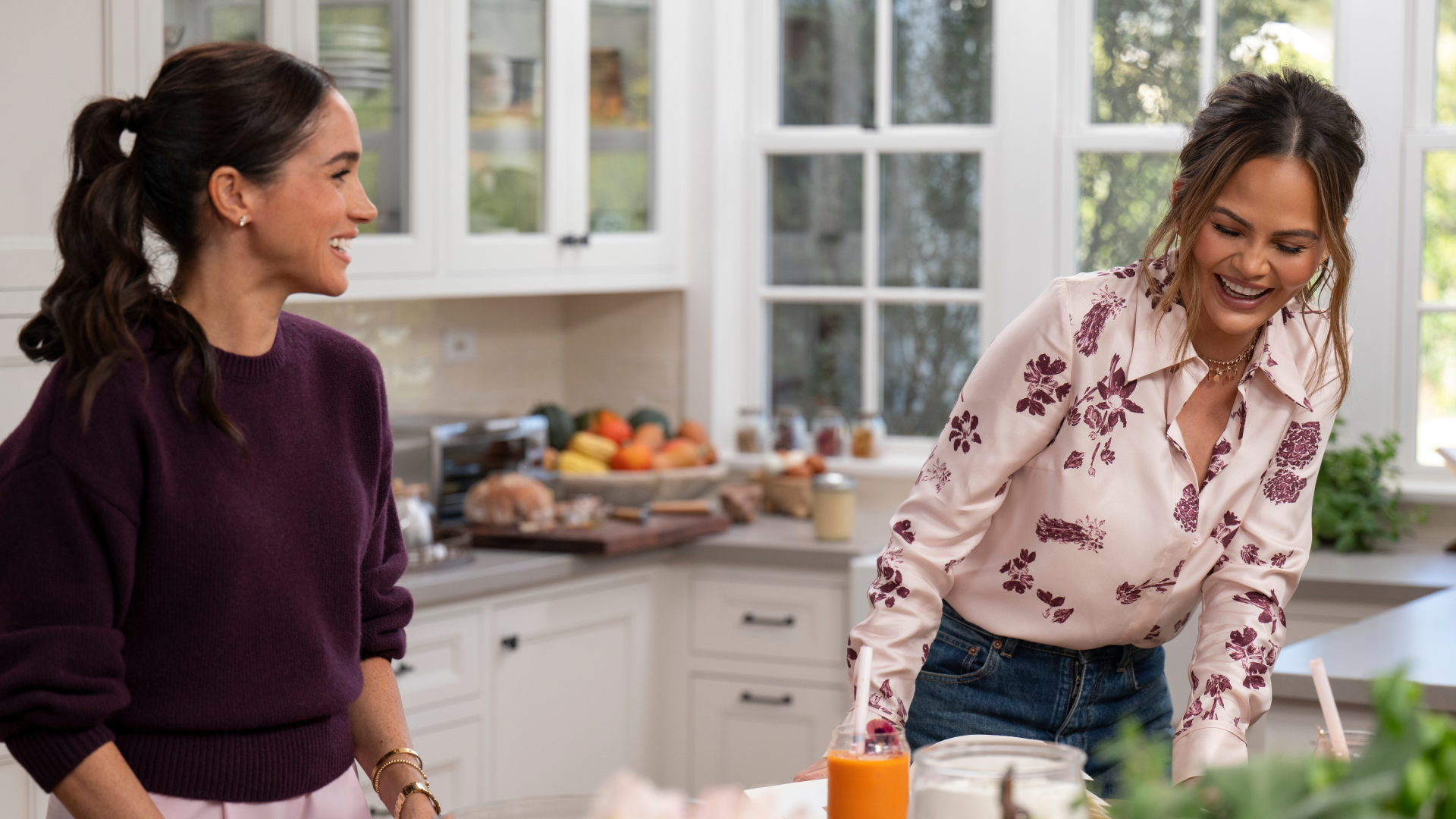 Meghan Markle wearing a maroon sweater looking at Chrissy Teigen, who is wearing a pink shirt laughing in a white kitchen