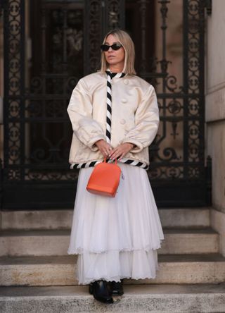 Karin Teigl is seen wearing black oval cat-eye sunglasses with a frame that is white at the top and shows the brand name from Chanel; a shiny cream-white silk jacket with oversized, black and white striped piping along the seams and large buttons from Louis Vuitton; multi-layered white mesh skirt with a ruffled hem from Louis Vuitton; a small orange leather “Alma” backpack with silver details carried by the handle from Louis Vuitton; a gold “Oura“ ring; three diamond rings; and black leather boots with a square toe and silver-detailed buckles from Billi Bi on November 05, 2024 in Vienna, Austria