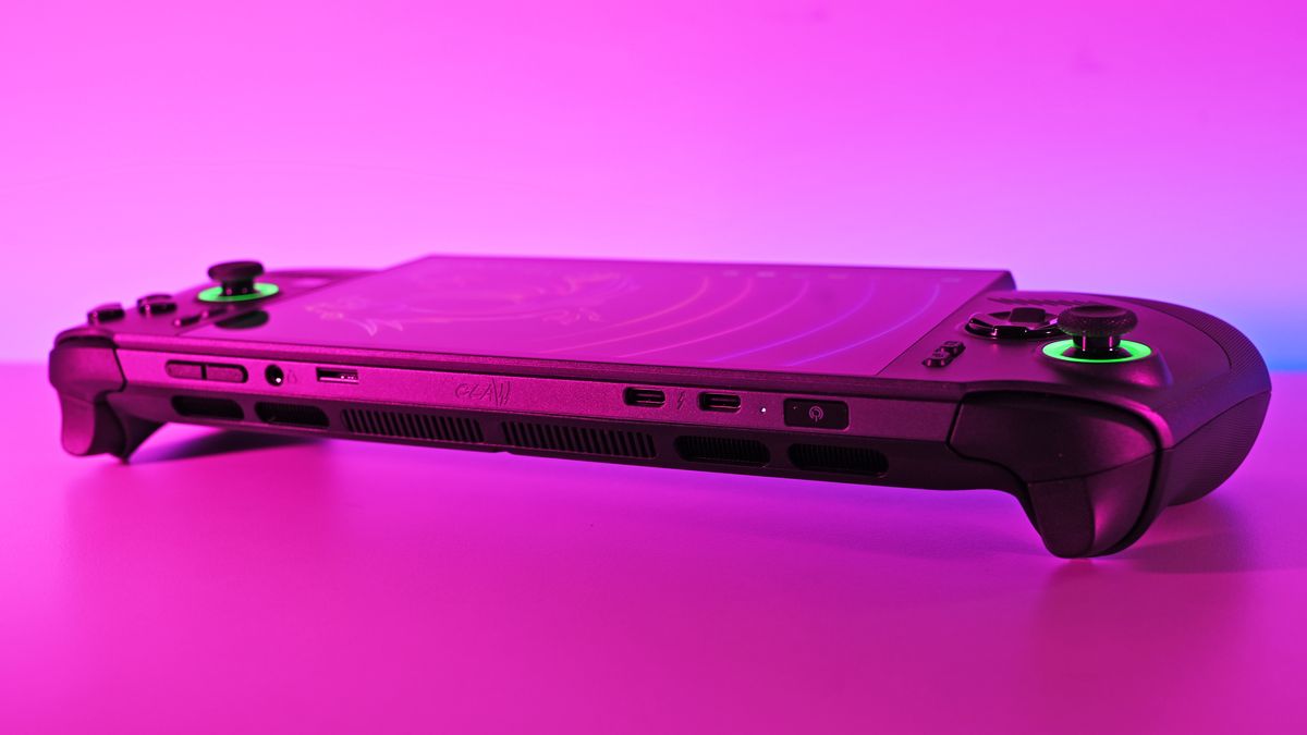 Wide shot of the console's top edge resting on a flat surface bathed in pink and blue neon light, showing the dual USB-C ports, power button, and glowing green joysticks.