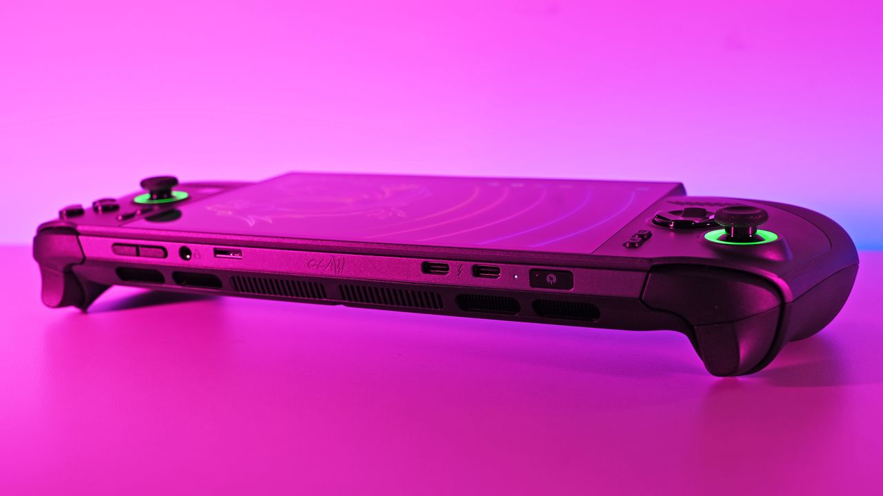 Wide shot of the console's top edge resting on a flat surface bathed in pink and blue neon light, showing the dual USB-C ports, power button, and glowing green joysticks.