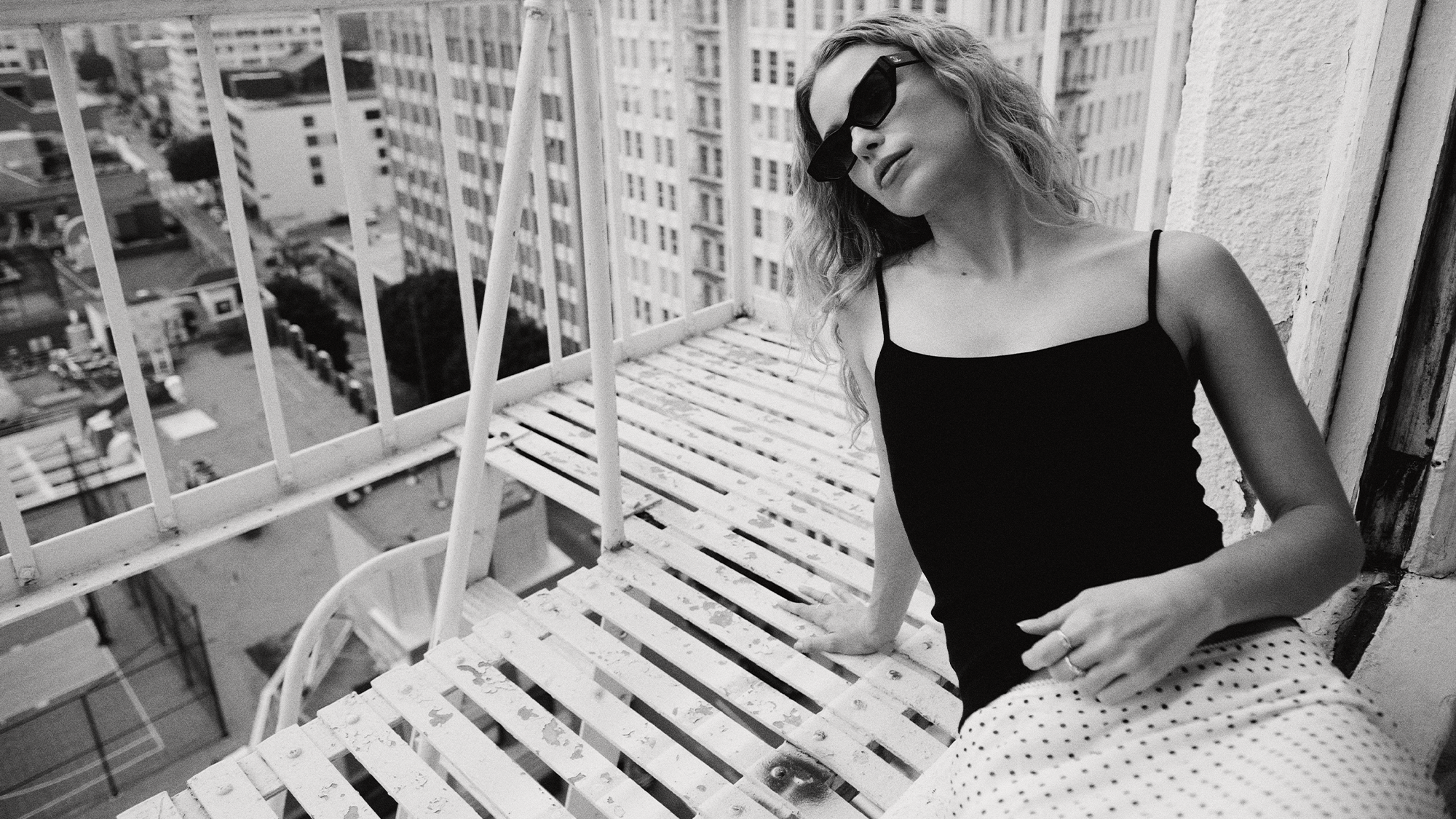 Elle Chapman leans out her window onto her NYC balcony wearing a black tank top and white and black polka dot skirt. She is wearing black sunglasses. 