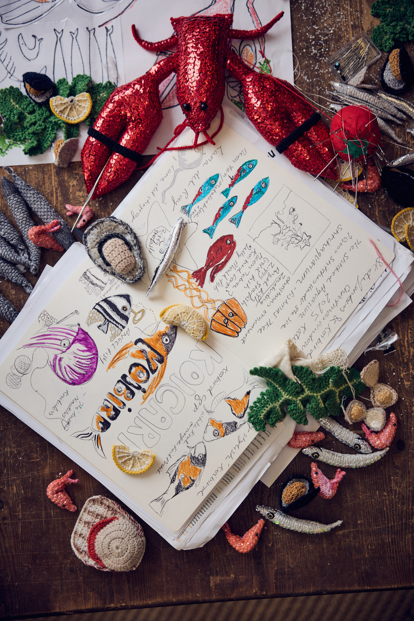 A sketched notebooks filled with fish-related drawings and notes sits atop a busy table with sardine-shaped objects, wreath fragments, lemon slices, and more drawings, as well as a toy lobster with glitters.