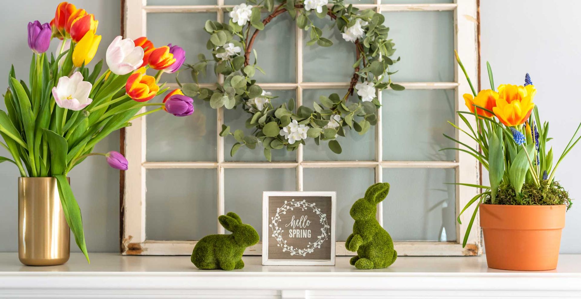 Easter mantel decor showing jugs of tulips, faux grass rabbits and a central Hello Spring sign