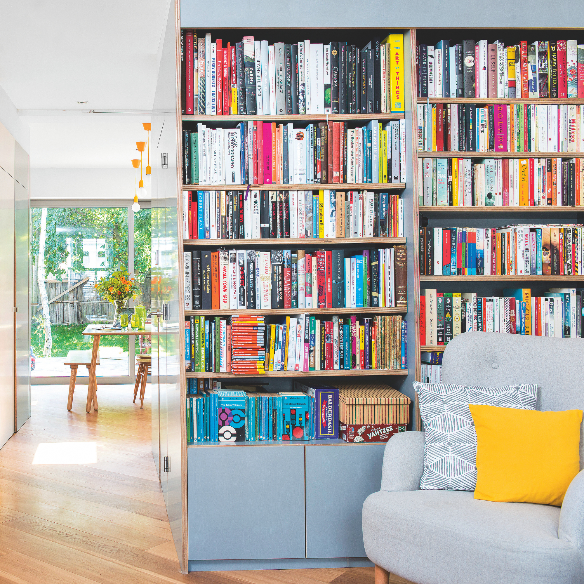Hallway with storage and bookcase and doors painted in duck egg blue, with staircase opposite with grey stair carpet and yellow and orange painted bannisters. 