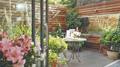 Metal bistro set on patio area in small enclosed garden surrounded by wooden panel walls and potted plants