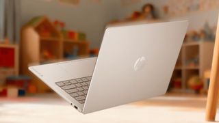 An open silver HP laptop sits on a table in a softly lit classroom, with shelves of toys and learning materials blurred behind it, creating a calm, welcoming atmosphere.