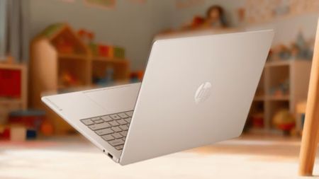 An open silver HP laptop sits on a table in a softly lit classroom, with shelves of toys and learning materials blurred behind it, creating a calm, welcoming atmosphere.