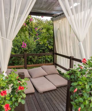 A dark brown wooden patio with brown floor pillows on it, a pergola with beige curtains, and green bushes with red and purple flowers on them