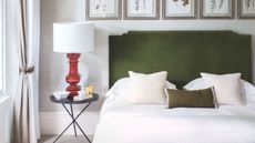 A neutral bedroom with a double bed with green velvet headboard, white bedding, black nightstand with red lamp and white lampshade, and five framed botanical prints above the bed.