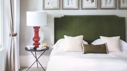 A neutral bedroom with a double bed with green velvet headboard, white bedding, black nightstand with red lamp and white lampshade, and five framed botanical prints above the bed.