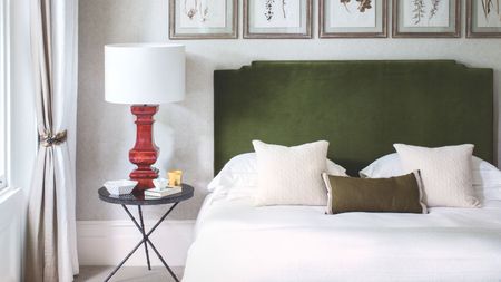 A neutral bedroom with a double bed with green velvet headboard, white bedding, black nightstand with red lamp and white lampshade, and five framed botanical prints above the bed.
