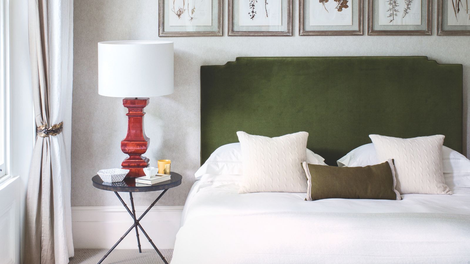 A neutral bedroom with a double bed with green velvet headboard, white bedding, black nightstand with red lamp and white lampshade, and five framed botanical prints above the bed.