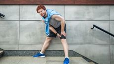 A man in running clothes pauses at the top of some steps, holding his knee and grimacing.