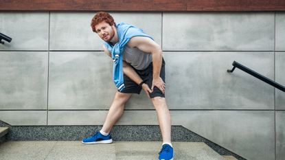 A man in running clothes pauses at the top of some steps, holding his knee and grimacing.