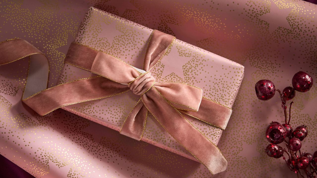 A gift wrapped in pink wrapping paper with gold dotted stars and a velvet ribbon beside a red berry twig