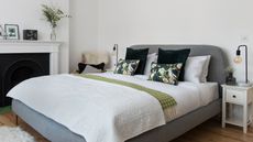 Grey upholstered double bed dressed with white bed linen in a white bedroom with warm wood floorboards and white bedside tables