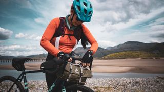 A woman leans over her bike's handlebars, adjusting the MSR Hubba Hubba Bikepack One-Person Tent attached to them in a green carry pouch.