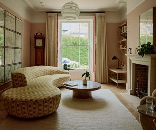 large soft pink painted living room with a floor to ceiling window and a large curved upholstered couch in front of a round coffee table and fireplace